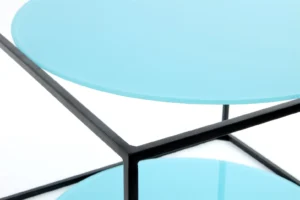 Close-up van de Bijzettafel Zwart, Blauw Rond Metaal, met het ronde lichtblauwe glazen blad en de plank met een geometrisch, eigentijds ontwerp en een minimalistisch zwart metalen frame.