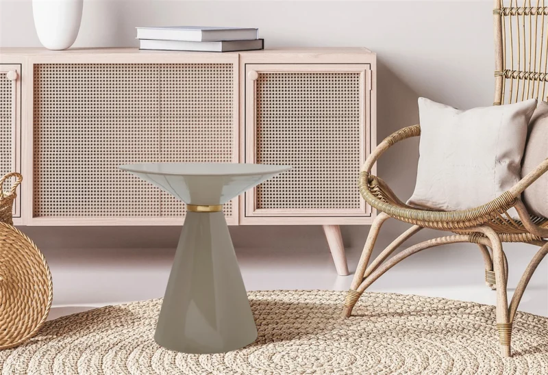 Een moderne woonkamer met een licht houten kast, een rotan stoel met een beige kussen, een rond geweven vloerkleed, een mand en de Bijzettafel Taupe Rond Ijzer - een klein rond ijzeren bijzettafeltje in taupe.