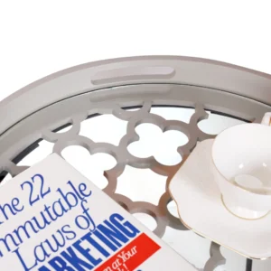 De Bijzettafel Grijs Metaal Design heeft een rond glazen blad met een stijlvol grijs metalen frame, dat een wit theekopje en schoteltje vasthoudt naast het boek "De 22 onveranderlijke wetten van marketing".