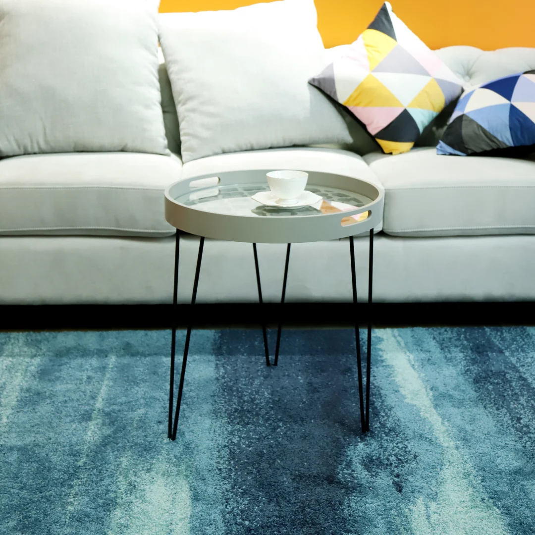 De Bijzettafel Grijs Metaal Design, een ronde grijze metalen tafel met dunne zwarte poten, staat op een blauw vloerkleed voor een lichtgrijze bank met kleurrijke geometrische kussens. Een witte kop en schotel rusten op de tafel.