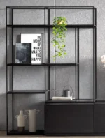 De Izola BOEKENREK DO, een strakke zwarte metalen plank, staat tegen een grijze muur met ingelijste kunst (op een ervan staat "DO MORE OF WHAT MAKES YOU HAPPY"), een hangende plant, gieter en decoratieve vazen.