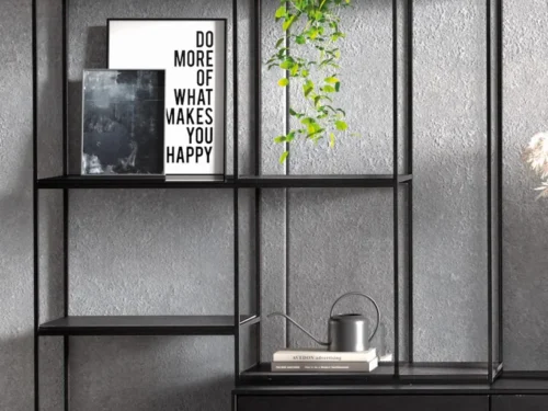 De Izola BOEKENREK DO, een strakke zwarte metalen plank, staat tegen een grijze muur met ingelijste kunst (op een ervan staat "DO MORE OF WHAT MAKES YOU HAPPY"), een hangende plant, gieter en decoratieve vazen.