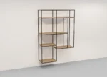 De Izola BOEKENREK HANG S04 is een minimalistisch wandrek met een zwart metalen frame en lichthouten planken in een asymmetrisch geometrisch ontwerp tegen een witte muur en grijze vloer.