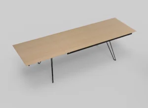 De Izola TAFEL 220-320 S04 is een lange, rechthoekige houten tafel met dunne, zwart metalen poten, tegen een effen lichtgrijze achtergrond. Het minimalistische ontwerp zorgt voor een moderne uitstraling.