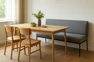 Een minimalistische eethoek met een houten tafel - perfect om te bespreken wat het verschil is tussen een salontafel en een bijzettafel - twee houten stoelen, een bank met grijze kussens, groene appels, een keramisch bord en een vaas met gebladerte bij het zonovergoten raam.