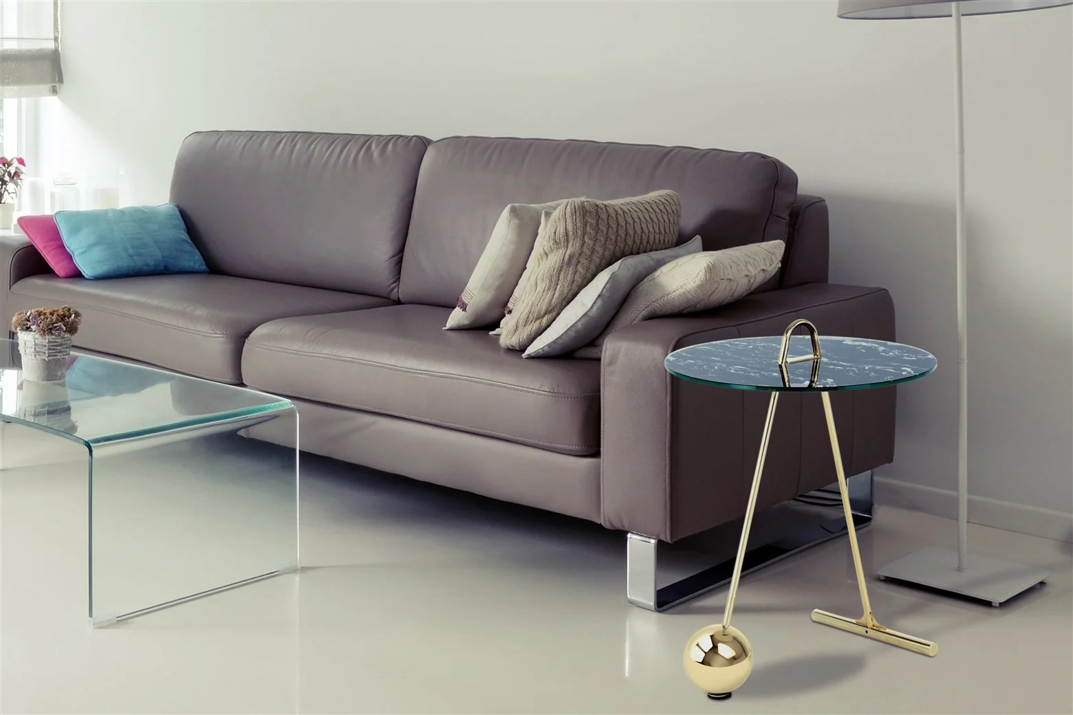 Een moderne woonkamer met een grijze leren bank met gemengde kussens, een salontafel van helder glas, een ronde Bijzettafel Zwart, Goud Staal bijzettafel en een hoge staande lamp. De inrichting is minimaal en eigentijds.