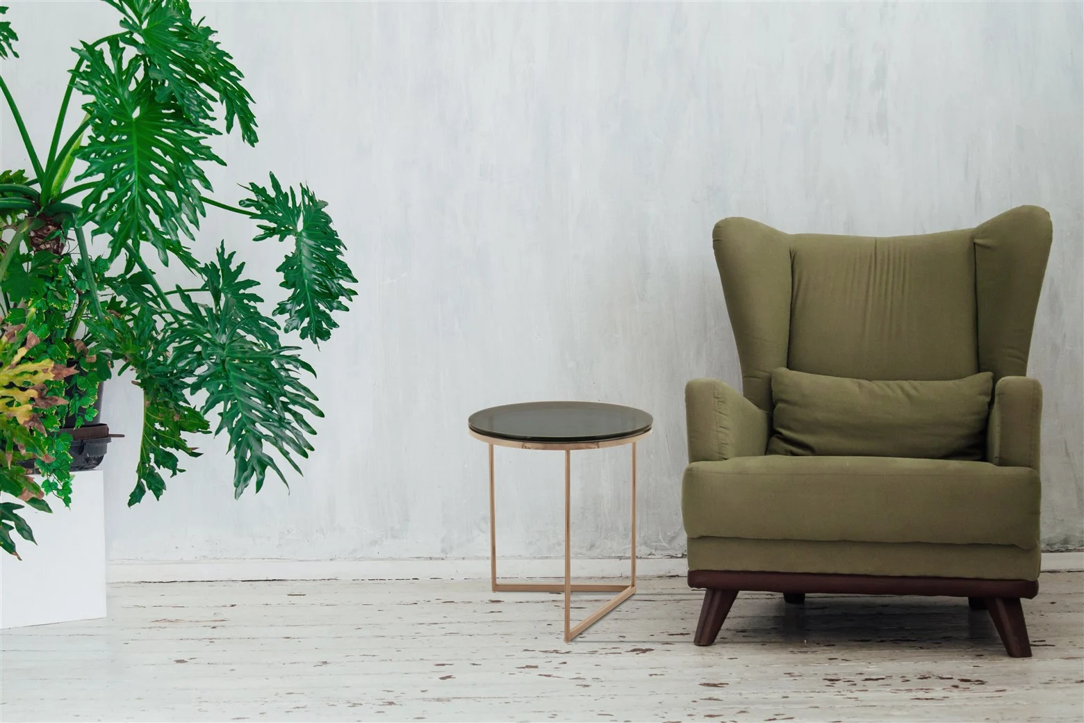 Een grote groene potplant staat naast de Bijzettafel Grijs Staal Design en een groen gestoffeerde fauteuil met kussen, allemaal tegen een verweerde witte muur en houten vloer.