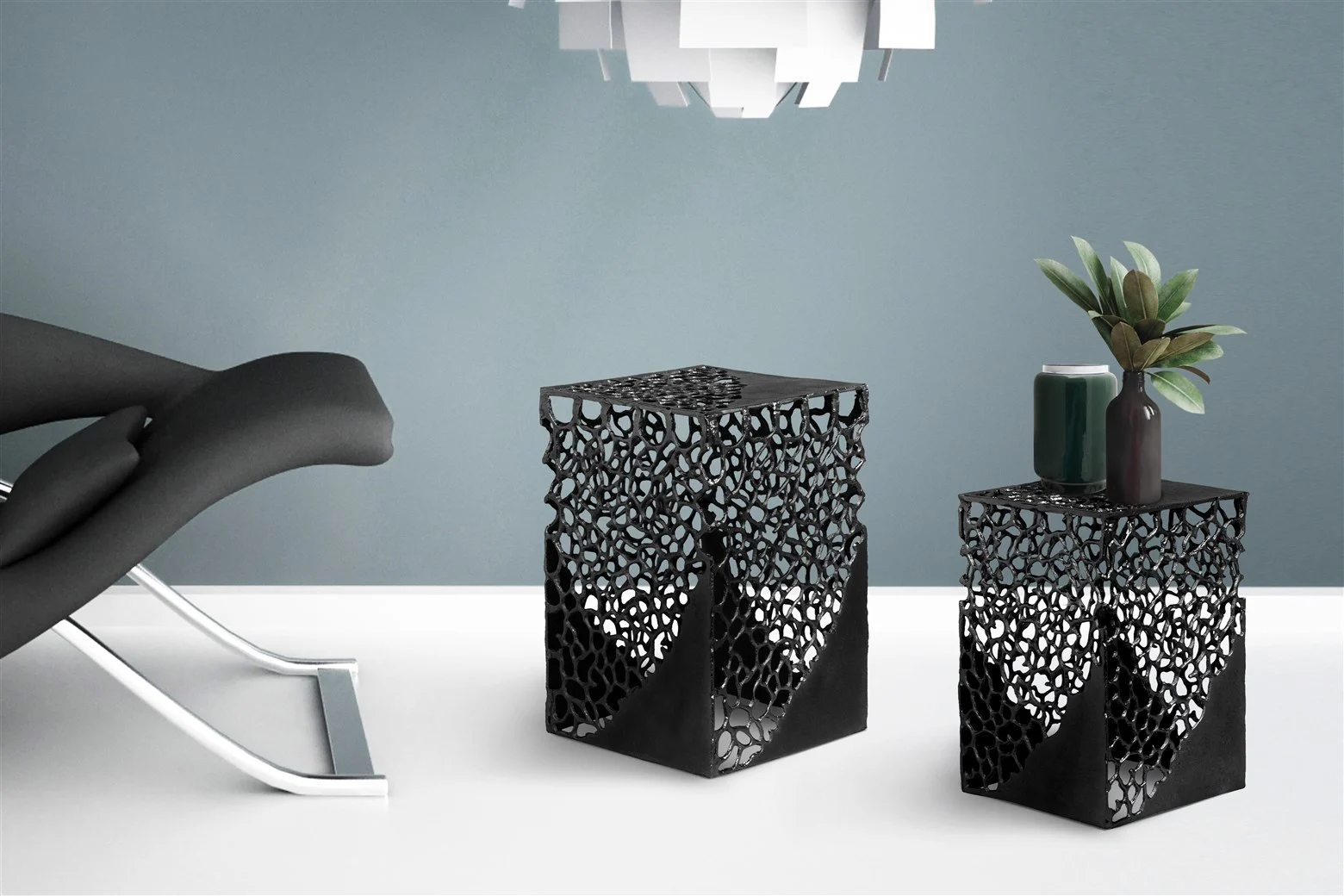 Een moderne minimalistische kamer met een zwarte loungestoel en twee Bijzettafels Zwart Aluminium Set. Op één tafeltje staan een potplant en een donker kopje. De muur is blauwgrijs en er hangt een witte hanglamp boven.