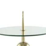 De Bijzettafel Transparant, Goud Staal is een moderne ronde glazen tafel met een centrale goudstalen steun met daarop een puntige punt en een ronde lus. Het metalen onderstel is gedeeltelijk zichtbaar.