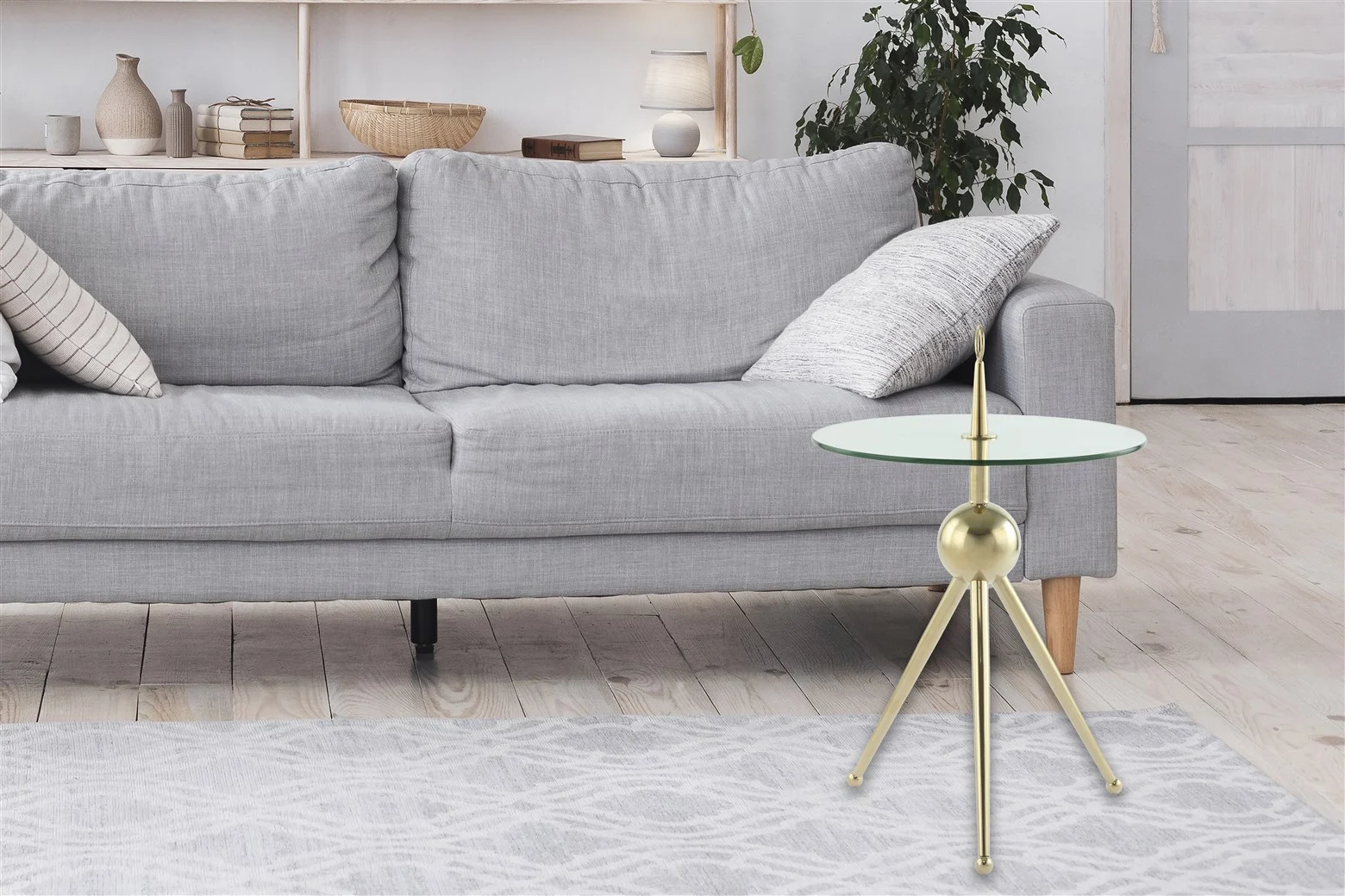 Een moderne woonkamer met een lichtgrijze bank, gedessineerde kussens, een Bijzettafel Transparant met goudkleurige stalen poten, een licht gedessineerd vloerkleed, een plant en met decor gevulde planken op de achtergrond.