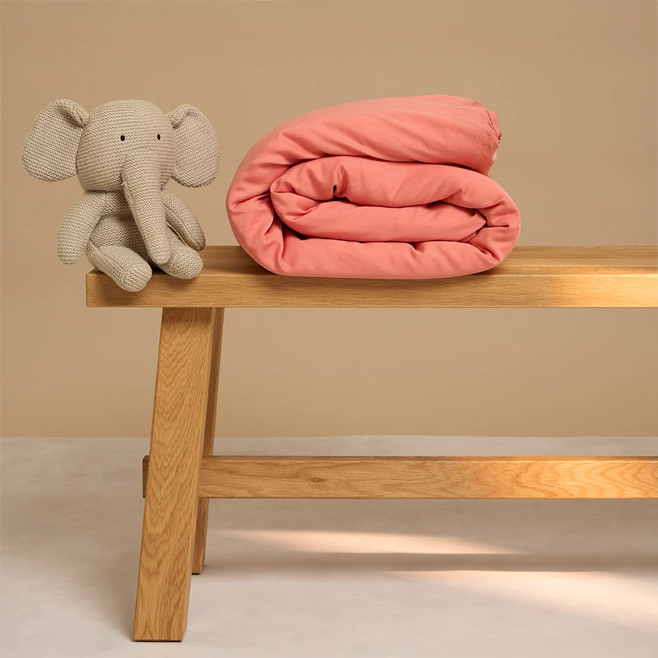 Een Dekbedovertrek 100x135 voor de slaapkamer is netjes opgevouwen en ligt naast een gebreide beige olifant op een houten bank met een beige achtergrond.