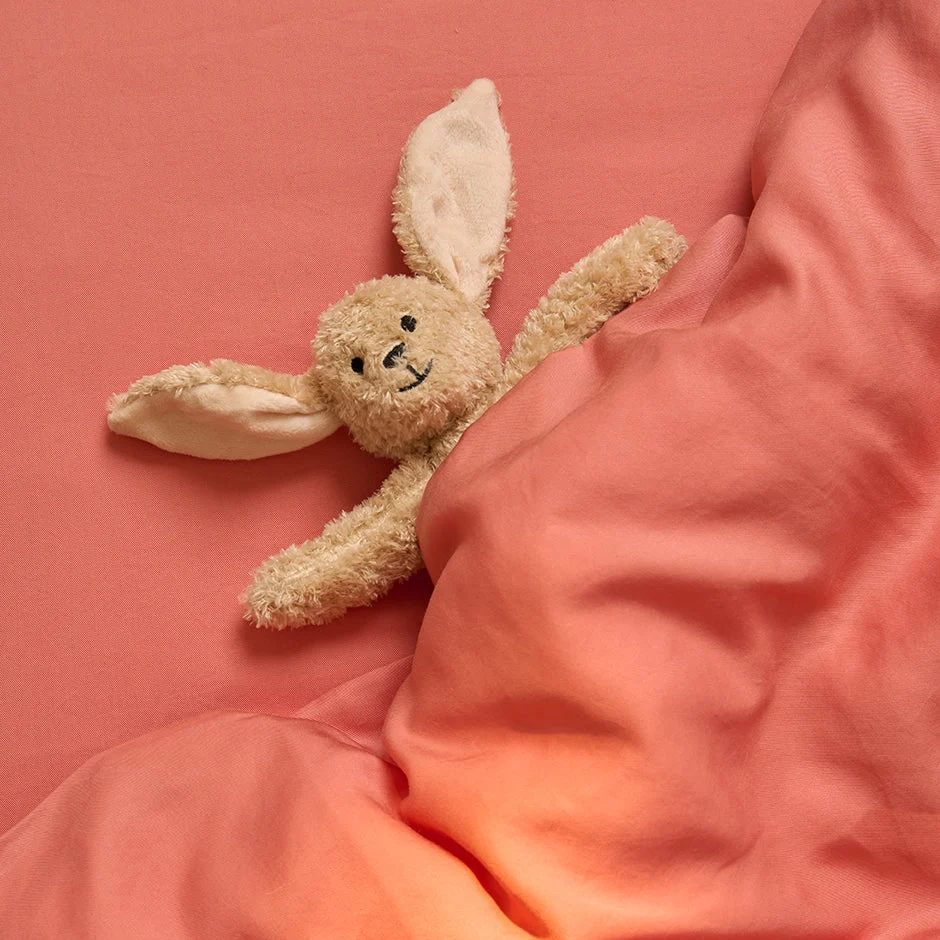 De Dekbedovertrek 100x135 voor de slaapkamer in koraalkleur bedekt het bed, met een pluchen konijntje met lange oren die onder de deken vandaan komen, zijn hoofd en armen zichtbaar.