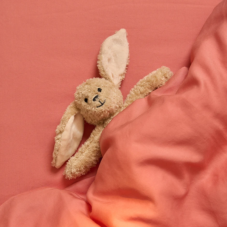 Een pluchen konijntje met lange oren ligt genesteld onder de Dekbedovertrek 100x135 voor de slaapkamer, rustend op een bijpassend roze laken en kijkt knus met een zachte glimlach.