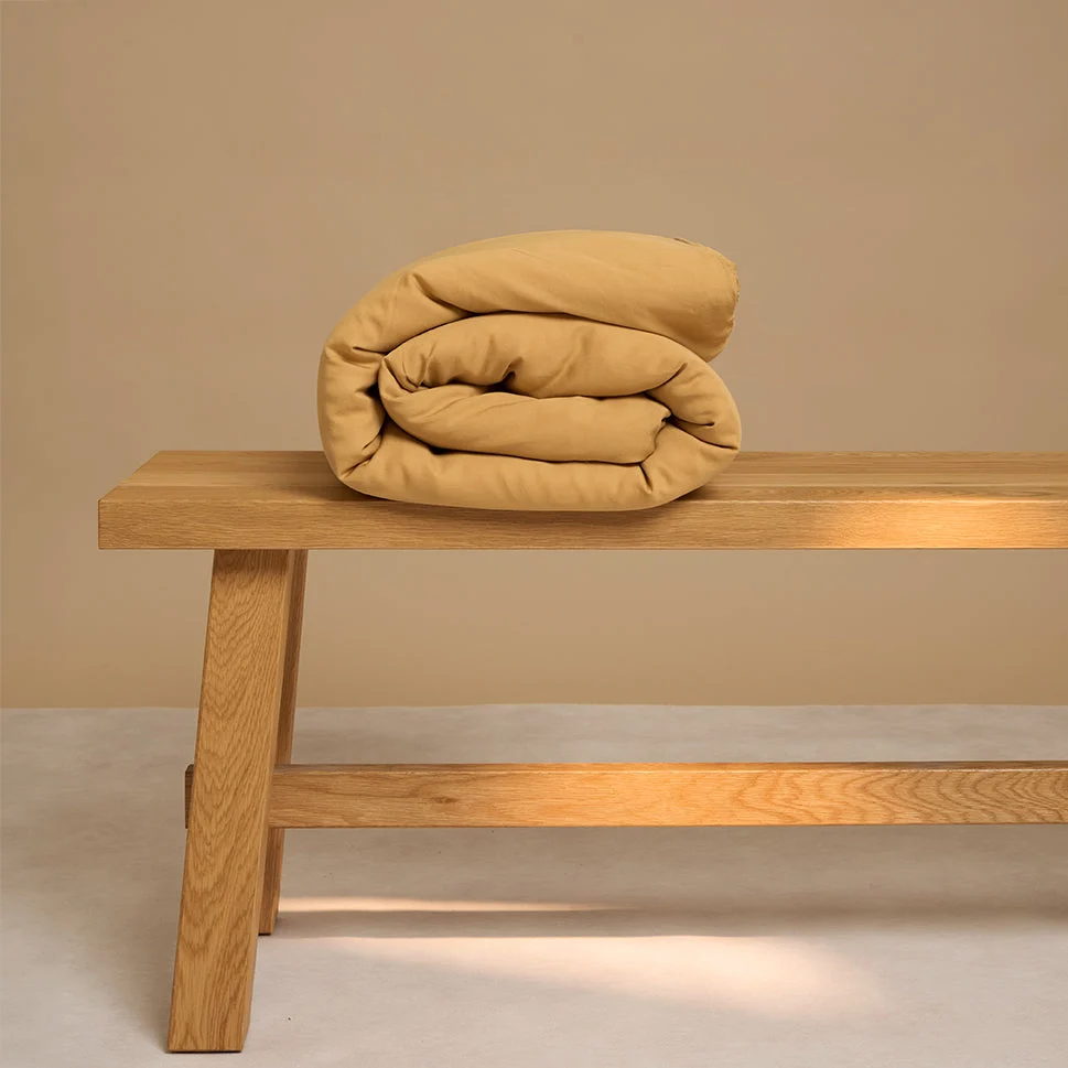 Een netjes opgerolde Dekbedovertrek Geel 100x135 voor de slaapkamer rust op de rechterkant van een lichthouten bank tegen een effen beige achtergrond.