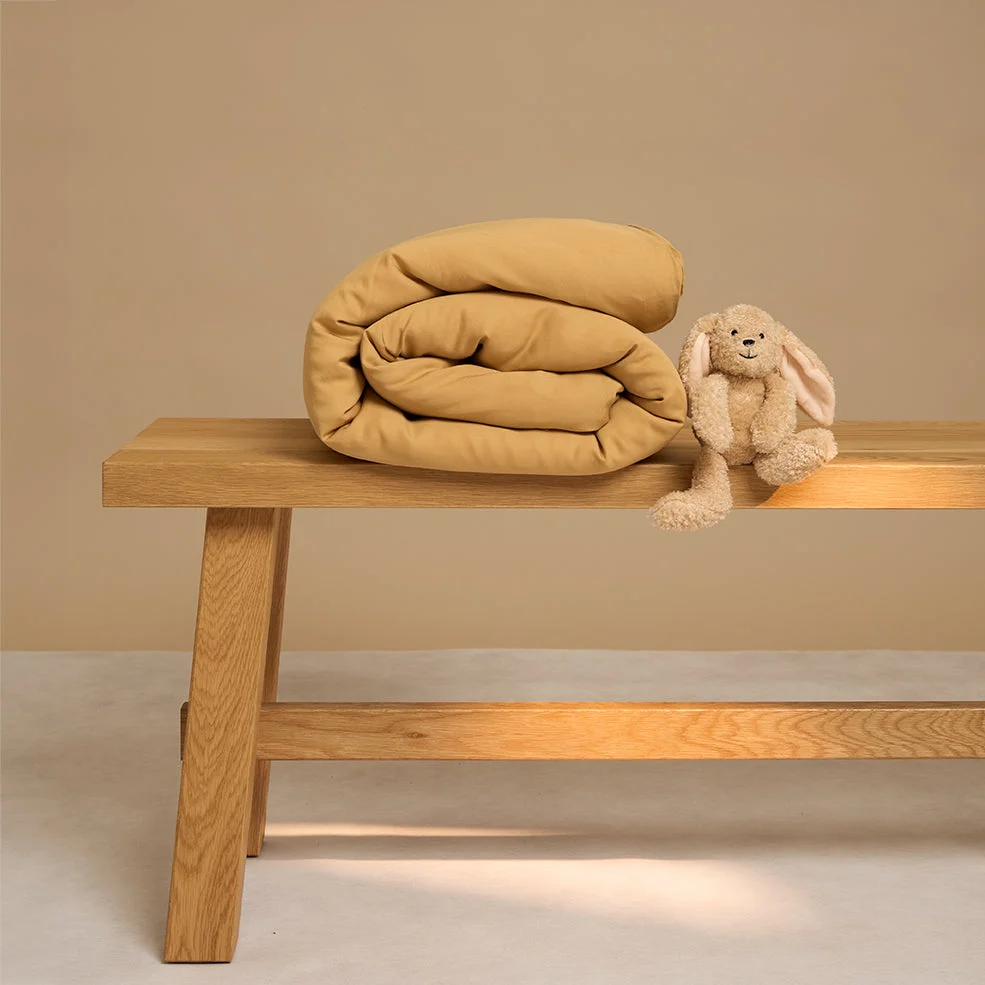 Een Dekbedovertrek 120x150 Geel voor elk seizoen ligt netjes opgerold op een houten bankje naast een zacht beige pluchen konijntje, alles tegen een neutrale achtergrond.