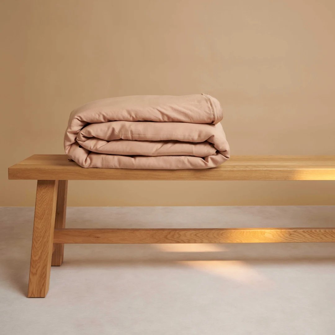 Gestapeld perzik dekbedovertrek op een houten bankje tegen een beige achtergrond. Zachte belichting benadrukt de textuur van het beddengoed.