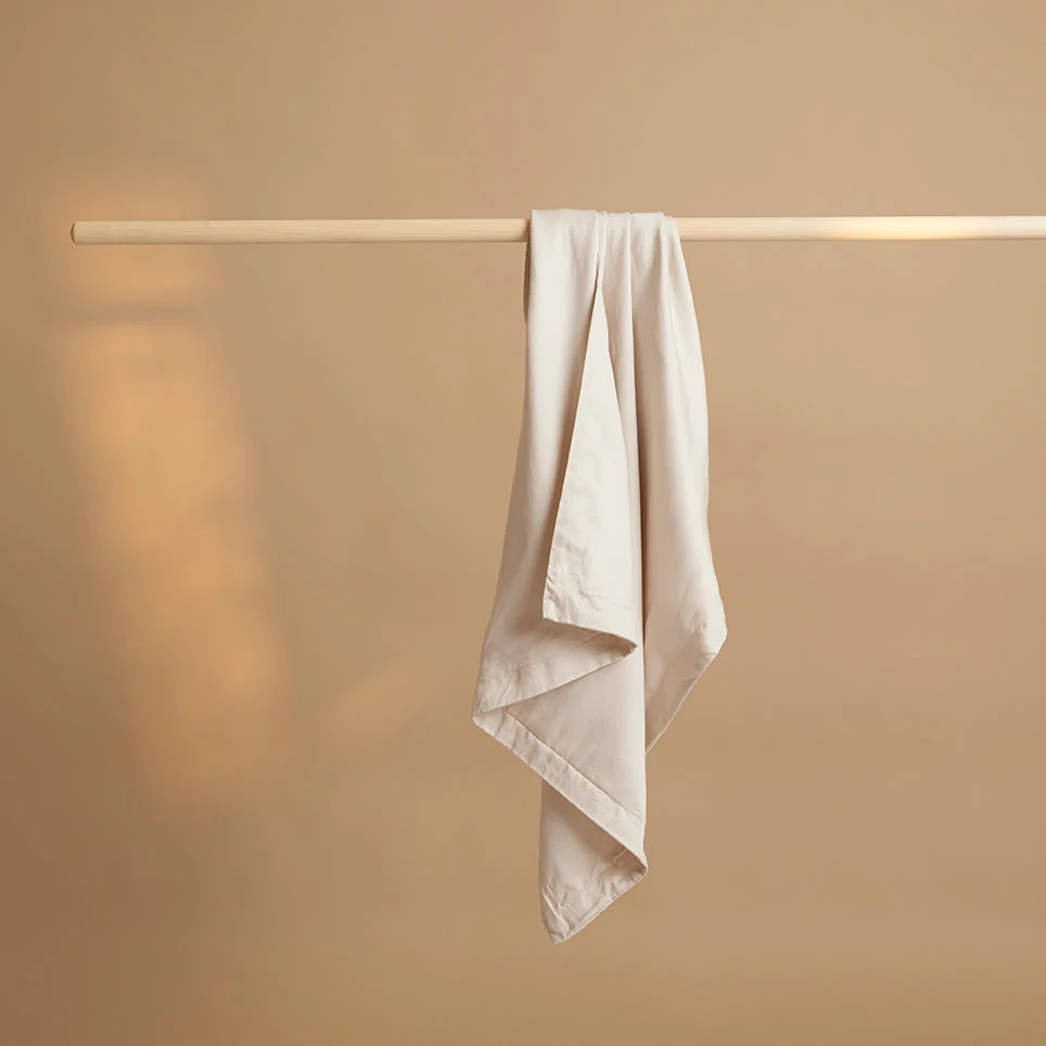 Beige kussensloop van TENCEL lyocell hangt over een houten stok. Zachte belichting, effen beige achtergrond. Strakke compositie, minimalistisch.