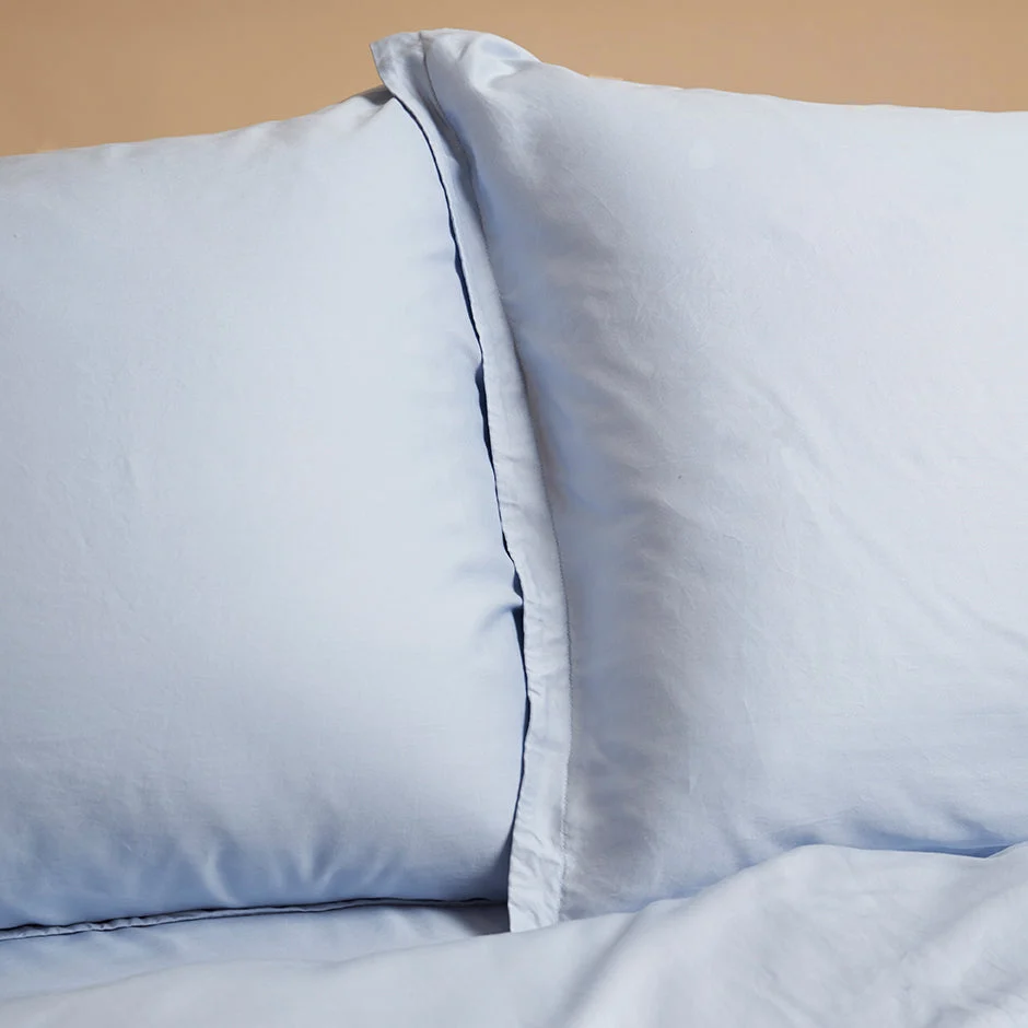 Detailopname van twee lichtblauwe kussenslopen van TENCEL lyocell op een bed. Zachte, egale belichting en een effen achtergrond.