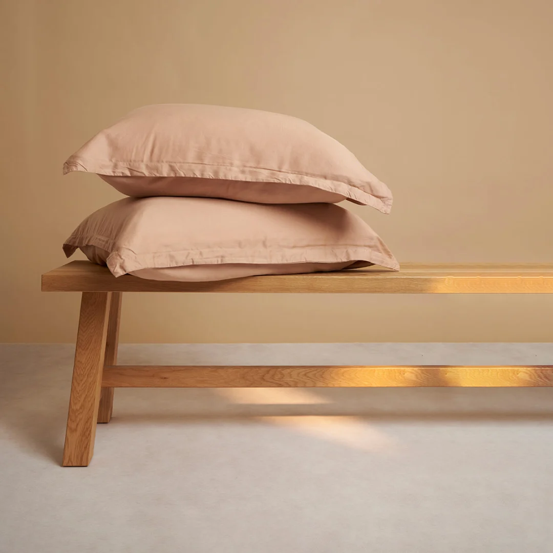 Twee perzikkleurige kussenslopen van TENCEL Lyocell op een houten bankje. Zachte kleuren en natuurlijk licht. Rustige en minimalistische compositie.