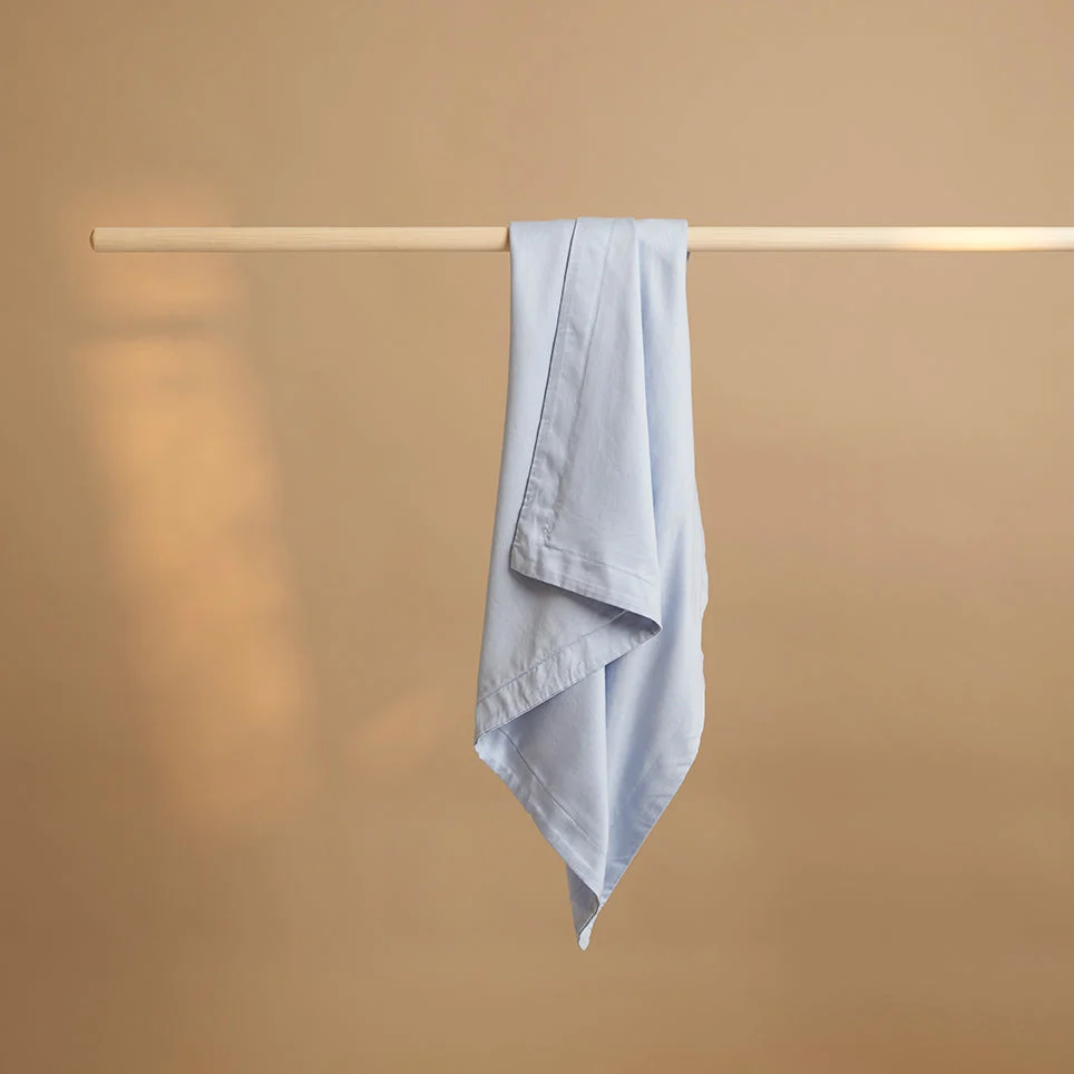 Lichtblauwe kussensloop van TENCEL lyocell hangt aan een houten stang. Zachte belichting, effen beige achtergrond. Close-up van de stof.