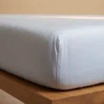 Detailopname van een lichtblauw TENCEL hoeslaken op een matras, strak getrokken. Zachte belichting benadrukt de textuur van de stof.