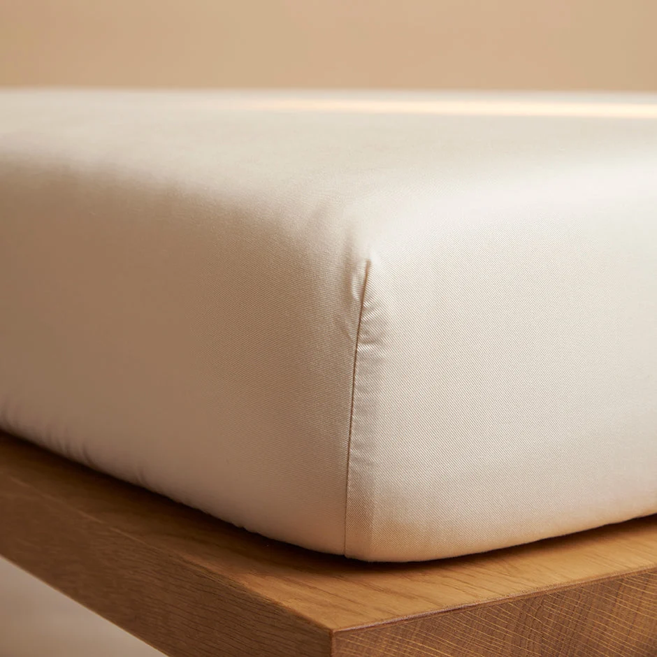 Beige hoeslaken detail op houten bed, zacht licht. Close-up van de strakke hoek, toont de kwaliteit en textuur van het TENCEL materiaal.