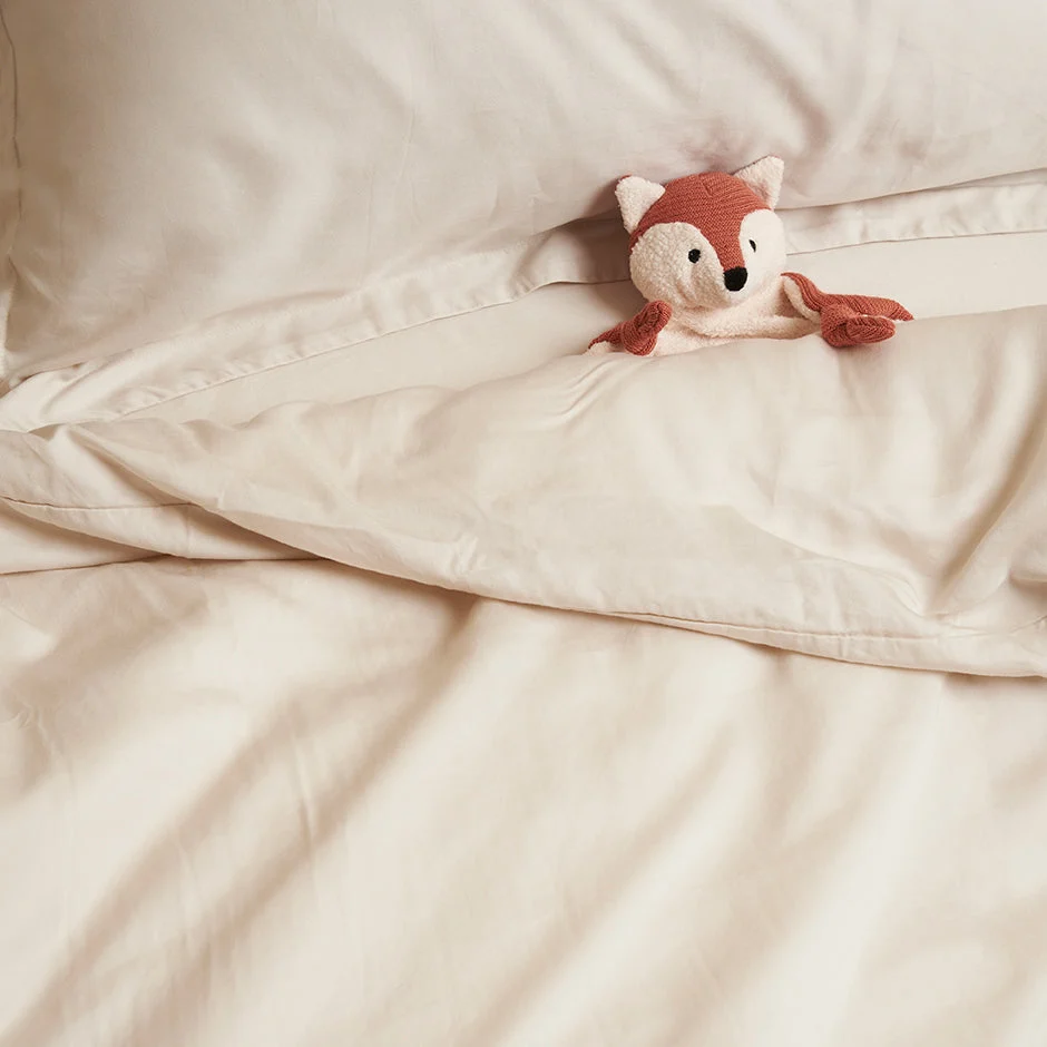 Beige hoeslaken van TENCEL™ twill stof, zacht en ademend. Een gebreide vos ligt op het beddengoed, zacht licht, close-up shot.