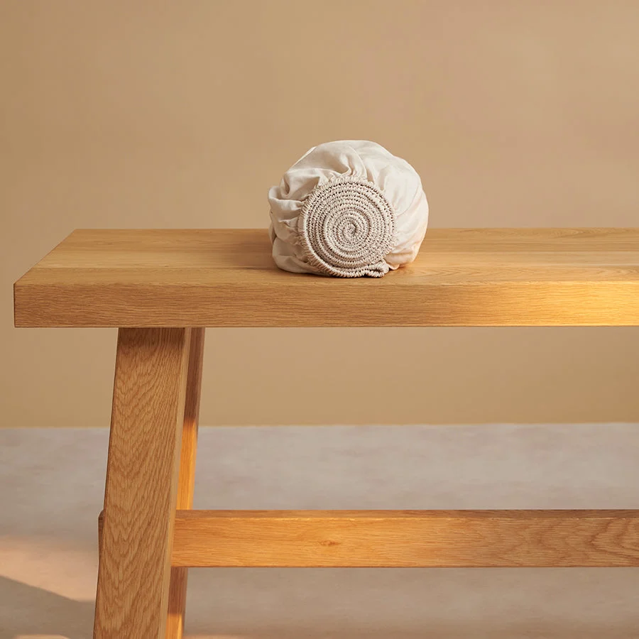 Beige ledikant hoeslaken, opgerold op een houten bankje. Zachte, warme kleuren en natuurlijk licht. Textuur van TENCEL™ Twill is zichtbaar.