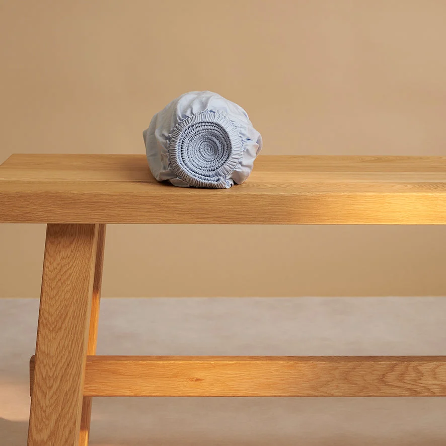 Lichtblauw hoeslaken van TENCEL lyocell op een houten bankje. Zachte kleuren en natuurlijk licht. De stof is opgerold en toont de elastische rand.