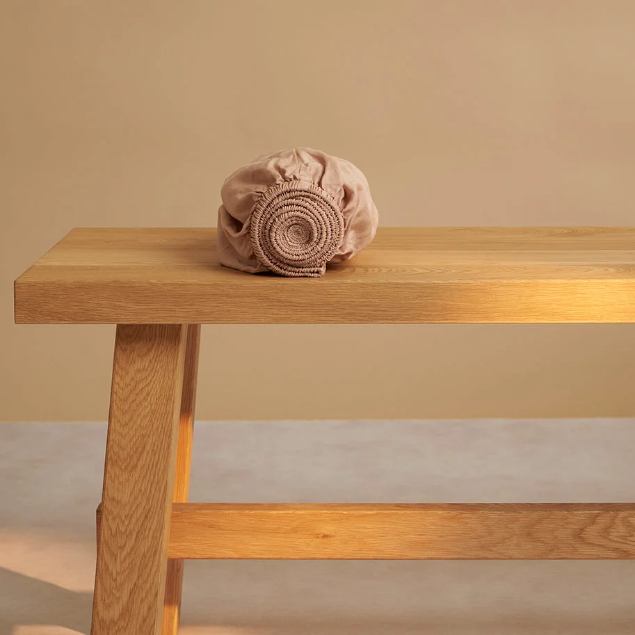 Peachkleurig hoeslaken van TENCEL™ twill op een houten bankje. Zachte kleuren, natuurlijk licht, focus op detail en textuur van het materiaal.