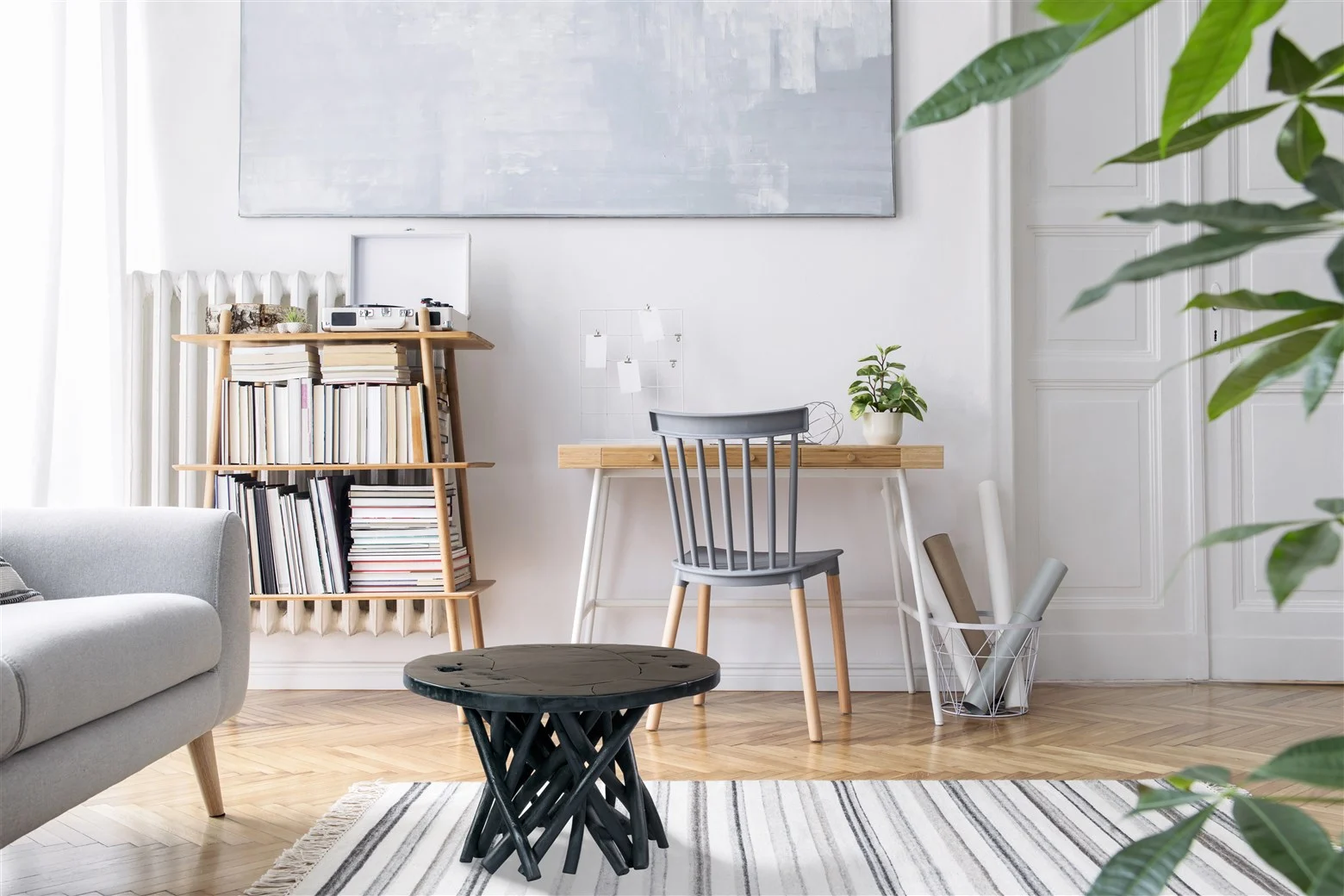 Een lichte, moderne werkkamer met een houten bureau, grijze stoel, de Bijzettafel Zwart Teakhout, boekenplank met boeken, muurkunst, planten in potten en een gestreept vloerkleed op een houten vloer.