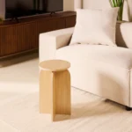 De Bijzettafel Felix - Naturel Ø45, een kleine ronde houten bijzettafel, staat op een licht vloerkleed naast een crèmekleurige bank met een wit kussen in een moderne woonkamer.