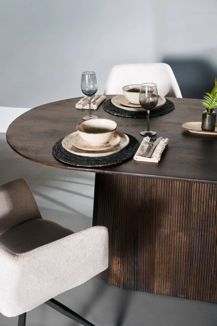 Een Mokka Eettafel Donkerbruin 180cm eetset voor twee personen, met beige gestoffeerde stoelen, neutrale borden en kommen, zwarte placemats, grijze wijnglazen, stoffen servetten en een klein plantaardig middenstuk in pot.