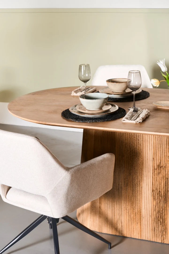 De Mokka Eettafel Lichtbruin 180cm is een ronde houten eettafel met twee beige stoelen met kussens, een set voor twee personen met borden, kommen, glazen en geweven placemats. Bovenop de tafel staat een witte bloem in een vaas tegen een lichtgroene muur.