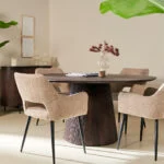 Een lichte, moderne eetkamer met de Mokka Ronde Eettafel Donkerbruin (Ø120cm), vier beige gestoffeerde stoelen, een vaas met rode bloemen, groene planten en minimalistische muurkunst in een neutrale omgeving.