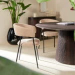 Een moderne eethoek met beige gestoffeerde stoelen, de Mokka Ronde Eettafel Donkerbruin (Ø120cm), grote groene planten en zachte natuurlijke verlichting.