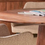 Een close-up van de Mokka Ronde Eettafel Lichtbruin - Ø150cm met een stapel tijdschriften erop, omringd door beige gestoffeerde stoelen.