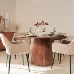 Een moderne eetkamer met de Mokka Ronde Eettafel Lichtbruin (Ø150cm), vier beige gestoffeerde stoelen, elegante couverts en roze bloemen in vazen. De ruimte is gestyled met neutrale muren voor een verfijnde look.