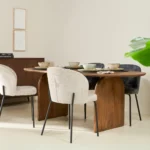 In een moderne eetkamer staat de Bella Eettafel Naturel - 180cm, een ovale houten tafel met vier stoelen met kussens - twee witte, twee donkergrijze - en een dressoir tegen een crèmekleurige muur. Een groen plantenblad verschijnt op de voorgrond.