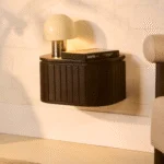 Een hand opent een verborgen lade in het Nachtkastje Donkerbruin Mangohout Industrieel zwevende nachtkastje, waardoor opbergruimte zichtbaar wordt onder een kleine lamp en boeken.