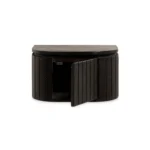 Het Nachtkastje is een compact, halfrond kastje gemaakt van donkerbruin mangohout met verticale groeven en een industriële look. Het heeft een enkele, gedeeltelijk openstaande deur die opbergruimte aan de binnenkant onthult.