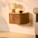 Het Nachtkastje Lichtbruin Mangohout, een industrieel houten zwevend nachtkastje met ribbel front, is gemonteerd aan een lichte wand. Bovenop staan een ronde lamp met glazen kap en een donker koffiekopje. Rechts is deels een beige bank zichtbaar.