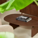 Een industrieel woonkamertafereel met de Salontafel 78 x 38 CM Mangohout ovale salontafel, een zwart-wit boek bovenop, beige gestoffeerde stoelen en groene bladeren die het uitzicht gedeeltelijk omlijsten.