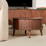 De Salontafel 65 CM Rond, een ronde houten salontafel met verticale groeven en vier poten, staat voor een houten mediaconsole naast een lichtgekleurde gestoffeerde stoel.