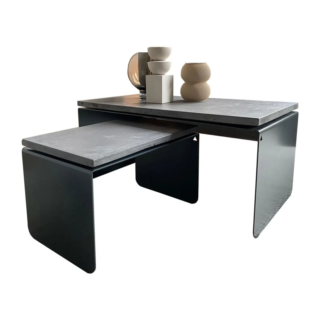 De Piramide Salontafelset heeft een modern ontwerp met een zwart metalen frame en bladen met betoneffect. Op de grotere tafel staan witte en beige vazen, terwijl de kleinere tafel er netjes gedeeltelijk onder staat.