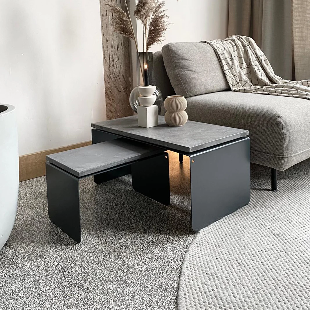 Een moderne woonkamer met een grijze fauteuil, gevouwen beige plaids en de Piramide Salontafelset met betonnen bladen, gestileerd met decoratieve vazen. Op de vloer ligt een grijs en crèmekleurig tapijt.