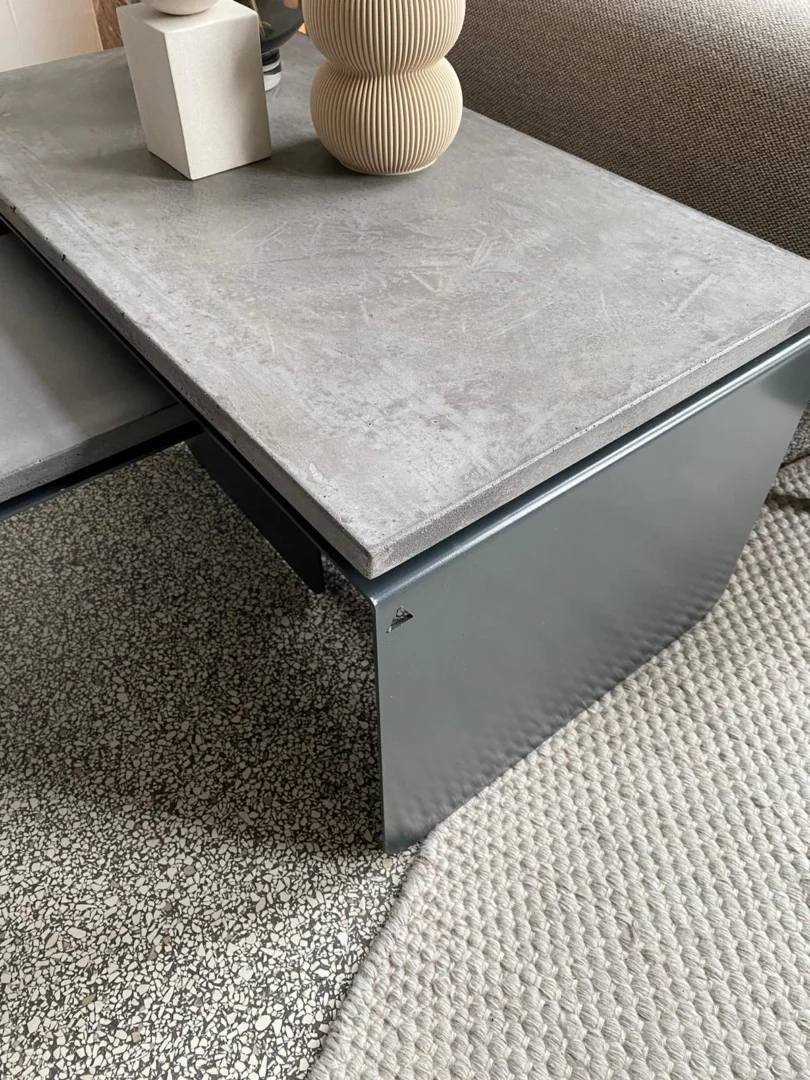 De Piramide - Salontafelset met een betonnen blad staat naast een bank op een geweven vloerkleed met een gespikkelde vloer, gestyled met een geribbelde beige vaas en een kleine witte kubus.