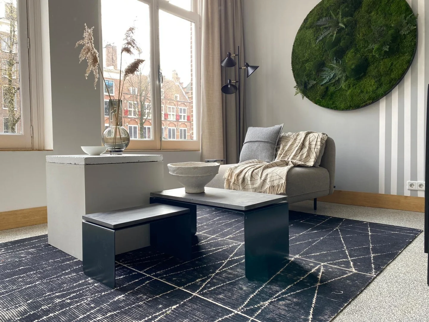 Een moderne woonkamer met een grijze fauteuil, een neutraal kleed, een Piramide salontafelset met betonnen blad, een zwarte bank, een geometrisch vloerkleed, een groot raam en ronde groene moskunst op een gestreepte accentmuur.
