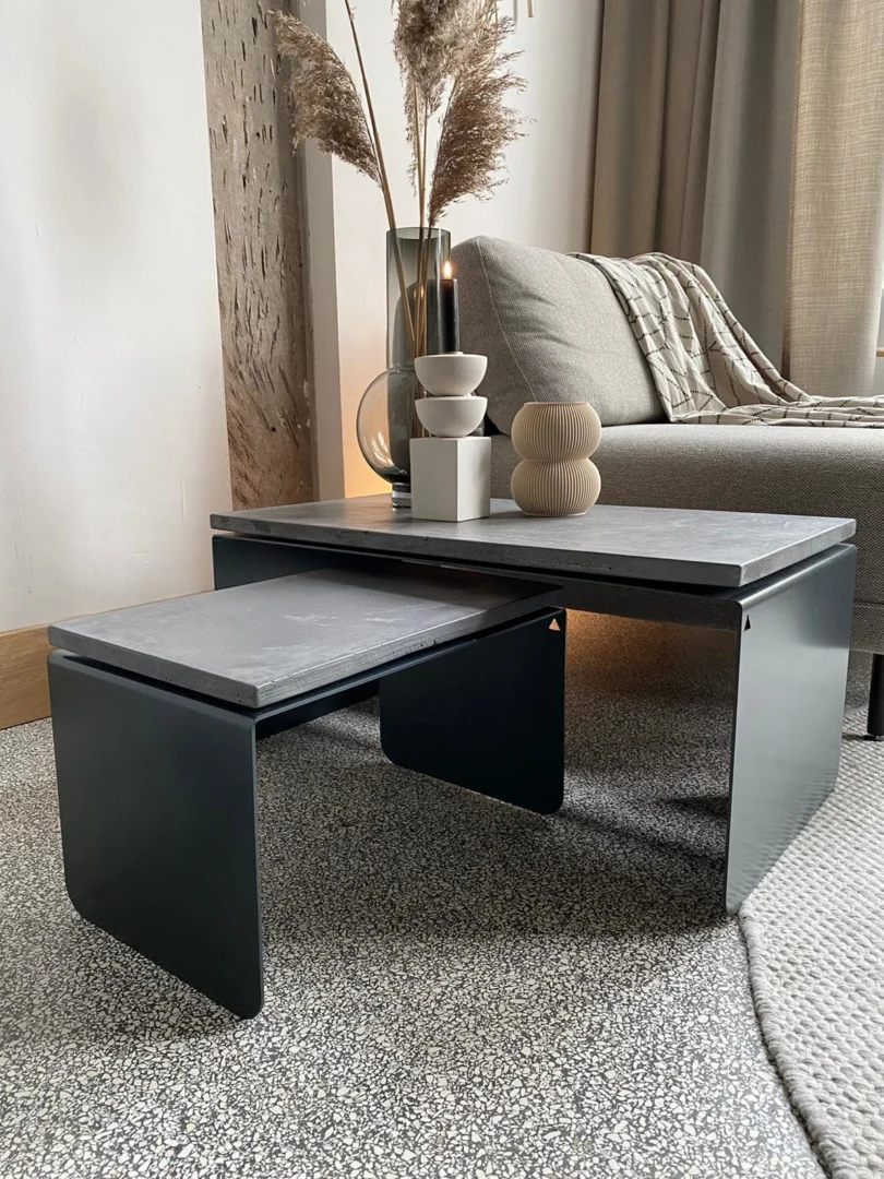 De Piramide Salontafelset met betonnen bladen en matzwarte poten staat op een gespikkeld tapijt in een gezellige woonkamer. Op de grotere tafel staan sculpturale vazen en pampagras, met een bank zichtbaar op de achtergrond.