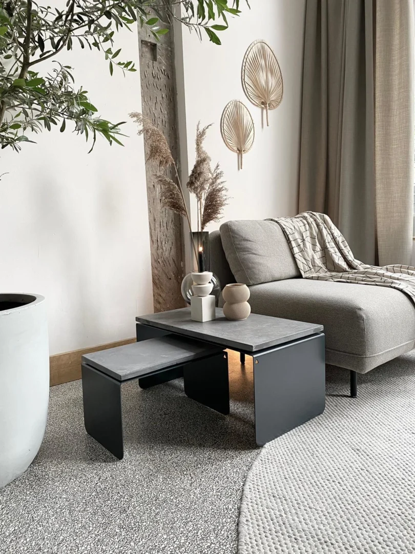 Een moderne woonkamer is voorzien van de Piramide Salontafelset met betonnen bladen, een grijze bank, neutraal gekleurde vazen en kopjes, een vloerkleed met structuur, een grote potplant en muurkunst met bladmotieven. Natuurlijk licht vult de ruimte.